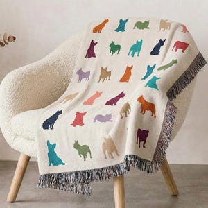 May include: A cream-colored throw blanket draped over a white chair. The blanket features a repeating pattern of colorful dog silhouettes in various poses. The blanket has a fringed edge.