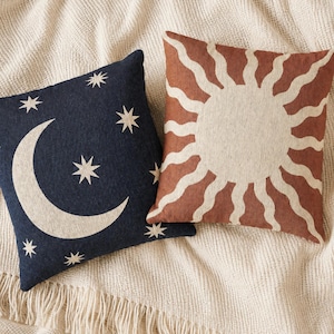 May include: Two square throw pillows with woven textures. One pillow is dark navy blue featuring a large cream-colored crescent moon and several eight-pointed stars. The second pillow is terracotta orange with a large cream-colored sun design.