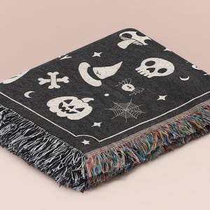 May include: A black woven throw blanket featuring a repeating white pattern of skulls, pumpkins, witch hats, spiders, spiderwebs, bones, stars, and crescent moons. The blanket has a decorative border and multicolored fringe along the edges.