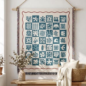 May include: A cream-colored wall tapestry with a grid pattern of blue and white sea-themed designs. The tapestry is framed with a red border and hangs from a wooden rod with a rope. A bench with a vase and basket is in front.
