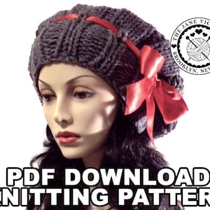 以下が含まれることがあります： 赤いサテンのリボンが付いた、濃い灰色のニット帽。帽子はリブ編みで、大きな赤いリボンで飾られています。「PDF DOWNLOAD KNITTING PATTERN」というテキストが画像の下部に表示されています。The Jane Victoria Brooklyn, New Yorkのロゴが右上隅にあります。
