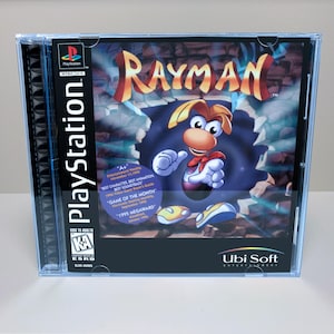 May include: A PlayStation video game case for Rayman featuring the title in large yellow and red letters. The cover art shows the character Rayman emerging from a dark portal against a stone background, with an ESRB Kids to Adults rating label.