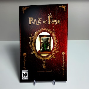 May include: A Rule of Rose software manual with a deep red, textured leather-like cover. The front features gold floral filigree surrounding a central oval window displaying a dark teddy bear on a chair. An ESRB Mature 17+ rating is in the corner.