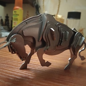 Puede incluir: Una escultura de toro de metal plateado con un diseño detallado y en capas. El toro está de pie sobre sus cuatro patas con la cabeza baja y los cuernos apuntando hacia adelante.