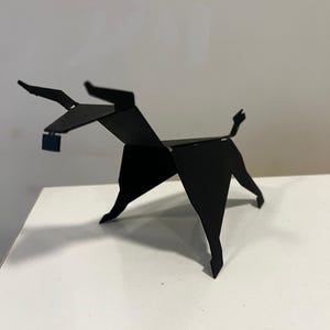 Op de afbeelding: Een zwarte origami sculptuur van een stier, staand op een wit oppervlak. De stier heeft hoorns, een staart en een klein vierkant object dat aan zijn nek hangt. De sculptuur is gemaakt van gevouwen papier of karton.