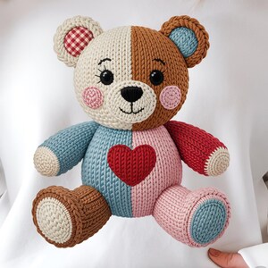 Peut inclure: Sweat-shirt blanc orné d'un motif d'ours en peluche crocheté. L'ours a un visage divisé, avec un côté marron et l'autre crème. Il a un cœur rouge sur la poitrine et des accents bleus, roses et rouges.
