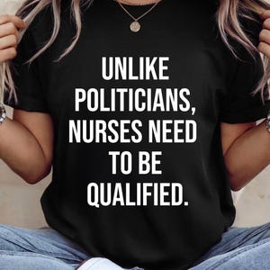Puede incluir: Camiseta negra con el texto blanco: "UNLIKE POLITICIANS, NURSES NEED TO BE QUALIFIED." La camiseta es de cuello redondo y la lleva una persona. El texto está en negrita.