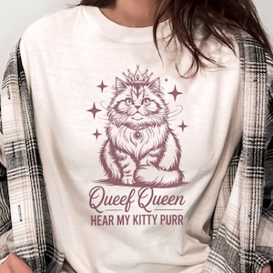 May include: Cream-colored t-shirt featuring a regal cat illustration wearing a crown, surrounded by stars. The text "Queef Queen Hear My Kitty Purr" is below the cat. The person is wearing a black and white plaid shirt.