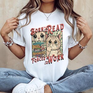 Peut inclure: T-shirt blanc avec un motif représentant un chat en larmes portant des lunettes et le texte "Born to Read, Pages Over Paychecks, Forced to Work". Le motif comprend une pile de livres et une esthétique vintage.