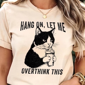 May include: A cream-coloured t-shirt featuring a black and white cat illustration drinking from a cup. The text above the cat reads "HANG ON, LET ME" and below the cat reads "OVERTHINK THIS". The shirt has short sleeves and a crew neck.