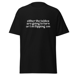 Puede incluir: Camiseta negra con el texto blanco "either the tables are going to turn or I'm flipping em". La camiseta es de estilo casual y manga corta.