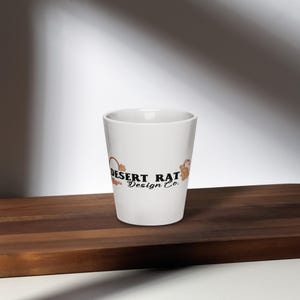 Könnte beinhalten: Ein weißer Keramik-Shot-Glas mit dem Text "Desert Rat Design Co." in Schwarz, mit einer braunen und orangefarbenen Grafik. Das Shot-Glas ist zylindrisch und steht auf einer dunkelbraunen Holzoberfläche.