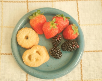 May include: A teal plate holds fresh strawberries, blackberries, and cookies. The strawberries are bright red with green stems. The blackberries are dark purple. The cookies are golden brown and shaped like pretzels and circles.
