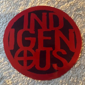 May include: A round sticker with the word "INDIGENOUS" in bold, red letters. The text is set against a dark background within a red circle. The sticker has a simple, bold design.