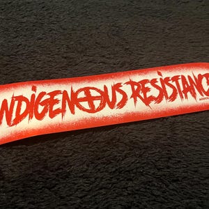 May include: A rectangular sticker with the words "INDIGENOUS RESISTANCE" in bold, red, brush-style lettering. The word "INDIGENOUS" features a symbol in place of the letter "O". The sticker has a white background with a red border and the text "WARPAINT SOCIETY".
