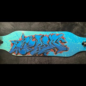 May include: A longboard skateboard with a blue and turquoise deck featuring a graffiti-style design in blue and red. The board has black wheels and trucks, and is designed for cruising and carving.