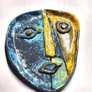 May include: A hand-sculpted ceramic dish shaped like an abstract face. The surface features a split color design with one side in deep blue and black tones and the other in bright yellow. The facial features are raised with defined eyes and lips.