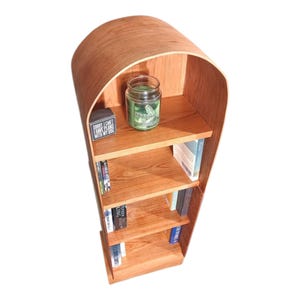 May include: A wooden bookshelf with a curved top and four shelves. The shelves hold books, a small jar, and a decorative block that reads "Sorry I can't have plans with my dog."