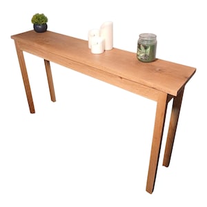 May include: A long, narrow wooden console table with a natural finish. The table has four angled legs and is topped with decorative items, including candles, a small potted plant, and a jar.