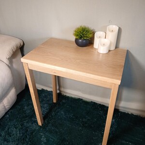 May include: A light-coloured wooden side table with four angled legs. The table has a small potted plant and three white pillar candles. The table is approximately 61 cm long and 41 cm wide.