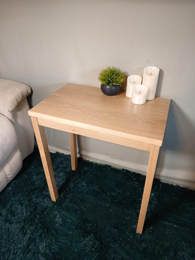 May include: A light-coloured wooden side table with four angled legs. The table has a small potted plant and three white pillar candles. The table is approximately 61 cm long and 41 cm wide.