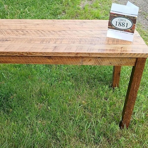 May include: A rectangular wooden bench with a natural brown finish and visible wood grain texture. A white card stands on the right side of the bench surface, featuring a dark oval emblem with the text CIRCA 1881 printed in black lettering.