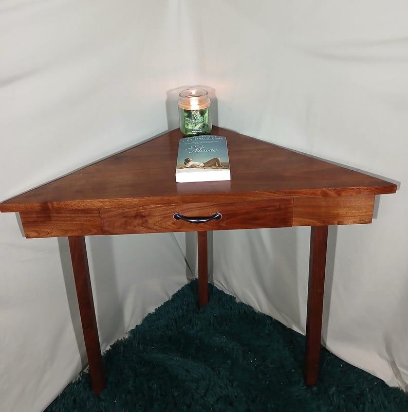 May include: A triangular wooden side table with a drawer and three legs. The table is a rich brown color and has a smooth, polished surface. A book and a candle in a glass jar sit on top. The table is approximately 30 inches wide.