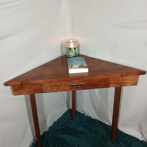 May include: A triangular wooden corner table with a drawer and dark brown legs. A book and a candle in a glass jar sit on the table. The table is made of a rich, dark wood.
