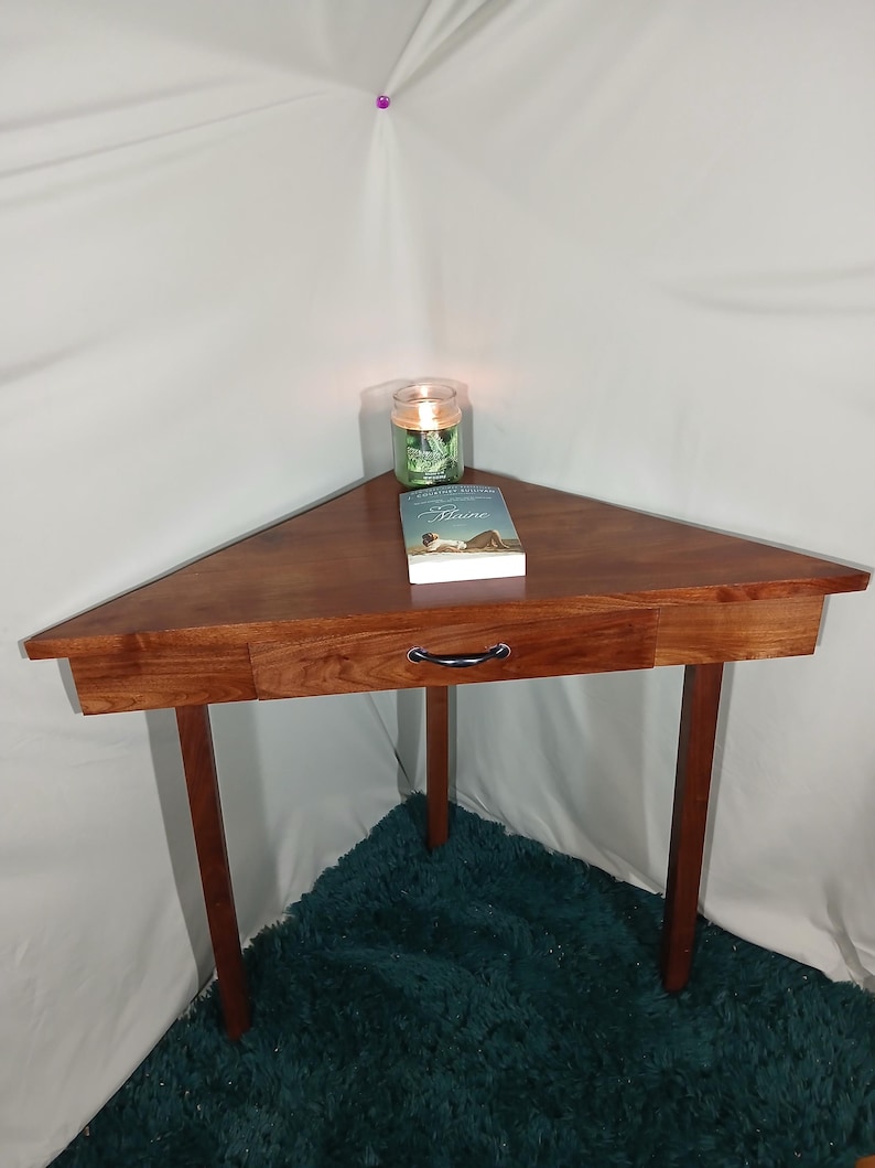 May include: A triangular wooden corner table with a drawer and dark brown legs. A book and a candle in a glass jar sit on the table. The table is made of a rich, dark wood.