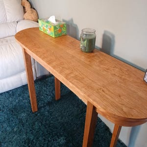 Puede incluir: Una consola de madera con acabado cerezo. La mesa tiene una tapa ovalada y cuatro patas delgadas. Una caja de pa&ntilde;uelos y una vela en tarro est&aacute;n sobre la mesa. La mesa est&aacute; sobre una alfombra color turquesa. La mesa mide unos 120 cm de largo.