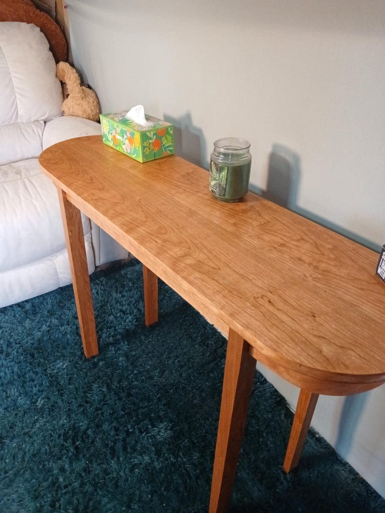 Puede incluir: Una consola de madera con acabado cerezo. La mesa tiene una tapa ovalada y cuatro patas delgadas. Una caja de pa&ntilde;uelos y una vela en tarro est&aacute;n sobre la mesa. La mesa est&aacute; sobre una alfombra color turquesa. La mesa mide unos 120 cm de largo.