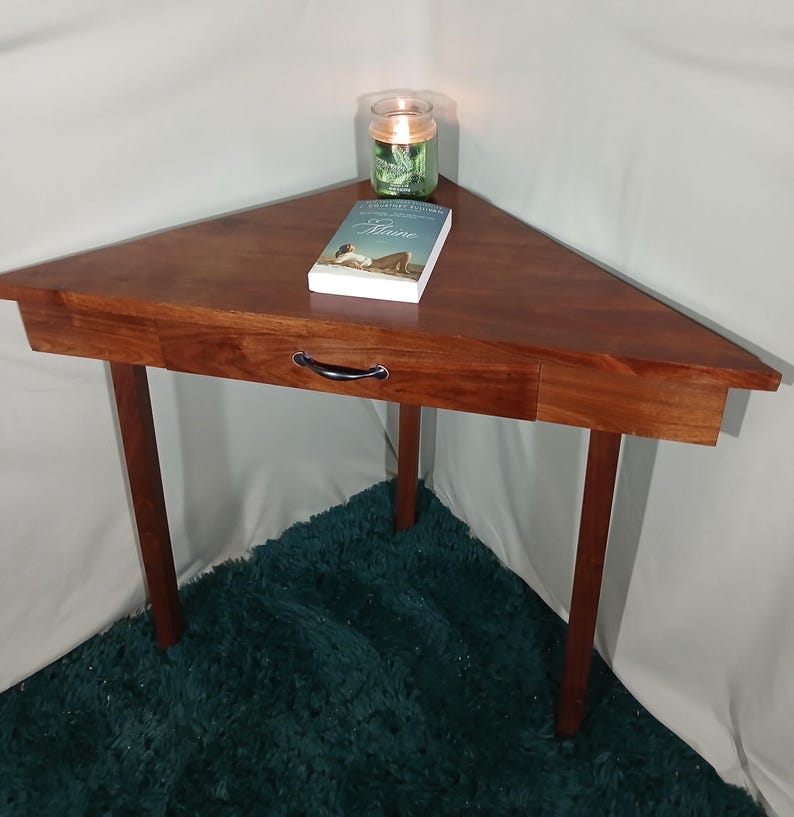 May include: A triangular wooden side table with a drawer and four legs. The table is a rich brown color and has a black handle on the drawer. A book and a candle in a glass jar sit on top of the table.
