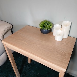 May include: A light-coloured wooden side table with a square top and four legs. The table is decorated with a small potted plant and three white pillar candles. The table is approximately 61 cm long and 61 cm wide.