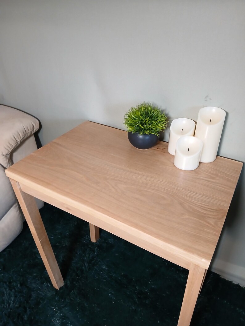 May include: A light-coloured wooden side table with a square top and four legs. The table is decorated with a small potted plant and three white pillar candles. The table is approximately 61 cm long and 61 cm wide.