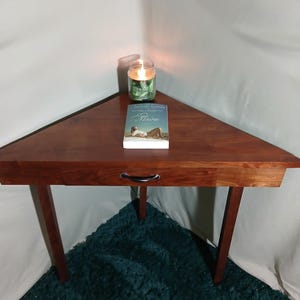 May include: A triangular wooden side table with a drawer and three legs. A lit candle in a glass jar and a book titled "Maine" are on the table. The table is brown, and the legs are dark brown.
