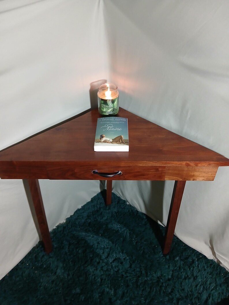 May include: A triangular wooden side table with a drawer and three legs. A lit candle in a glass jar and a book titled "Maine" are on the table. The table is brown, and the legs are dark brown.
