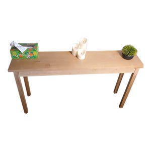 May include: A light-colored wooden console table with a rectangular top and four angled legs. The table is decorated with a tissue box, three white candles, and a small potted plant. The table is approximately 48 inches long.
