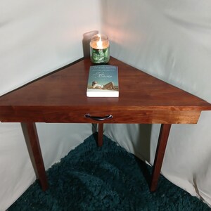 May include: A triangular wooden side table with a drawer, featuring a rich, dark brown finish. A book titled "Maine" and a lit green candle sit on top. The table has three legs and is placed on a teal rug.