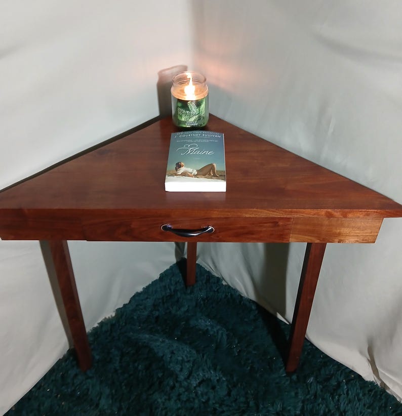 May include: A triangular wooden side table with a drawer, featuring a rich, dark brown finish. A book titled "Maine" and a lit green candle sit on top. The table has three legs and is placed on a teal rug.
