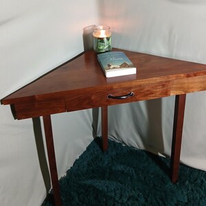 May include: A triangular wooden side table with a drawer and black handle. A book and a lit candle sit on the table's surface. The table has four legs and is made of dark wood. The table is approximately 30 inches wide.