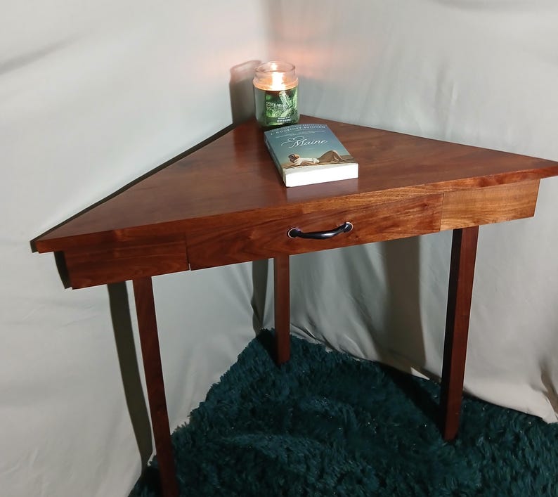 May include: A triangular wooden side table with a drawer and black handle. A book and a lit candle sit on the table's surface. The table has four legs and is made of dark wood. The table is approximately 30 inches wide.