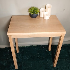 May include: A light-coloured wooden side table with a square top and four straight legs. The table is decorated with a small potted plant and three white pillar candles. The table is approximately 61 cm wide and 61 cm long.