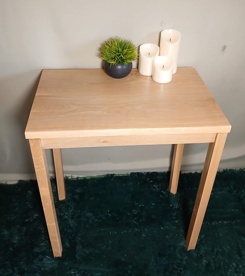 May include: A light-coloured wooden side table with a square top and four straight legs. The table is decorated with a small potted plant and three white pillar candles. The table is approximately 61 cm wide and 61 cm long.