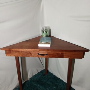 May include: A triangular wooden side table with a drawer and black handle. A glass jar and a book are on the table. The table has four legs and is made of dark wood.