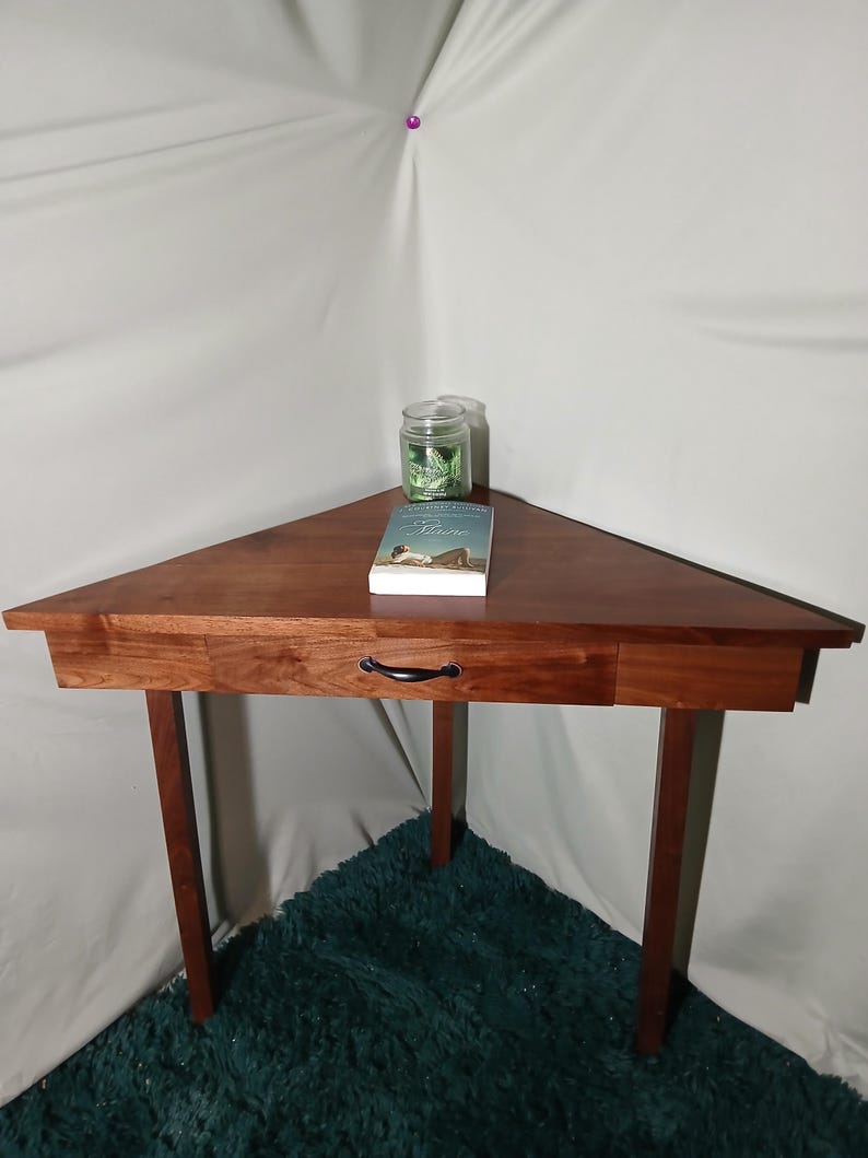 May include: A triangular wooden side table with a drawer and black handle. A glass jar and a book are on the table. The table has four legs and is made of dark wood.