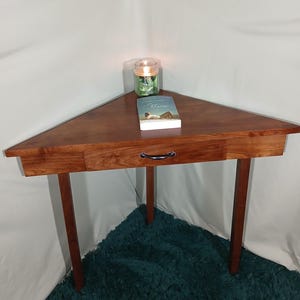 May include: A triangular wooden corner table with a drawer and three legs. A book and a lit candle in a glass jar sit on the table. The table is approximately 30 inches wide.