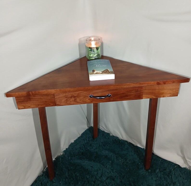 May include: A triangular wooden corner table with a drawer and three legs. A book and a lit candle in a glass jar sit on the table. The table is approximately 30 inches wide.