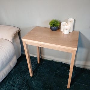 May include: A light oak side table with a rectangular top and four angled legs. The table has a small potted plant and three white pillar candles. The table is next to a bed. The table is approximately 60 cm in height.