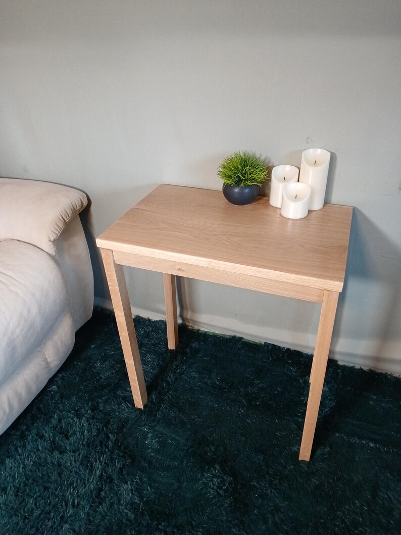 May include: A light oak side table with a rectangular top and four angled legs. The table has a small potted plant and three white pillar candles. The table is next to a bed. The table is approximately 60 cm in height.
