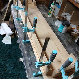 May include: A woodworking project with a long wooden plank held together by several blue and black clamps. The clamps are securing the wood on a dark surface. Tools and supplies are visible in the background.